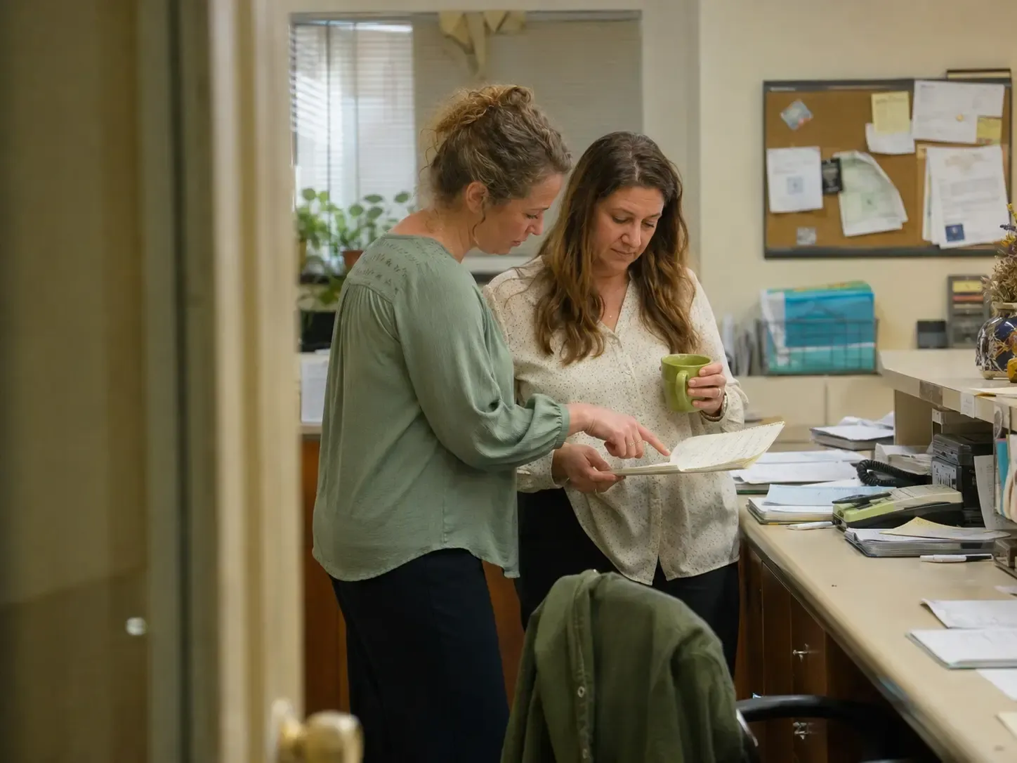 Two senior living admissions coworkers comparing handwritten call notes in a busy back office