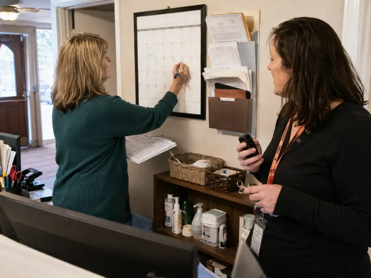 Two community staff coordinating a tour change with a wall calendar, phone, and keys